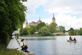 Eine Fotografie des Burgsees zeigt am linken Ufer sitzend eine Person in historischer schwarz-weißer Uniform. Neben ihr steht eine Frau in einem historischen Kleid mit weitem Reifrock, gefedertem Hut und Sonnenschirm aus Spitzenstoff. Sie winkt zwei Personen auf dem Burgsee in einem kleinen Ruderboot. Diese tragen ebenfalls historische Kleidung. Im Hintergrund ist hinter Bäumen das Schweriner Schloss.