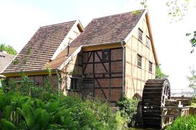  Rechts von dem dreigeschossigen Fachwerkhaus befindet sich ein Mühlrad aus Holz. Die Schleifmühle spiegelt sich in dem bewuchterten Mühlbach.
